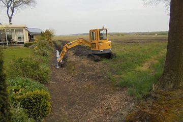 Singelonderhoud met een gele graafmachine langs de beplanting naast een tuin.