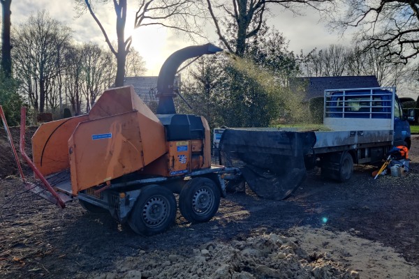 Oranje houtversnipperaar in actie met uitgeworpen snippers, naast een vrachtwagen van Groentechniek Klomp.